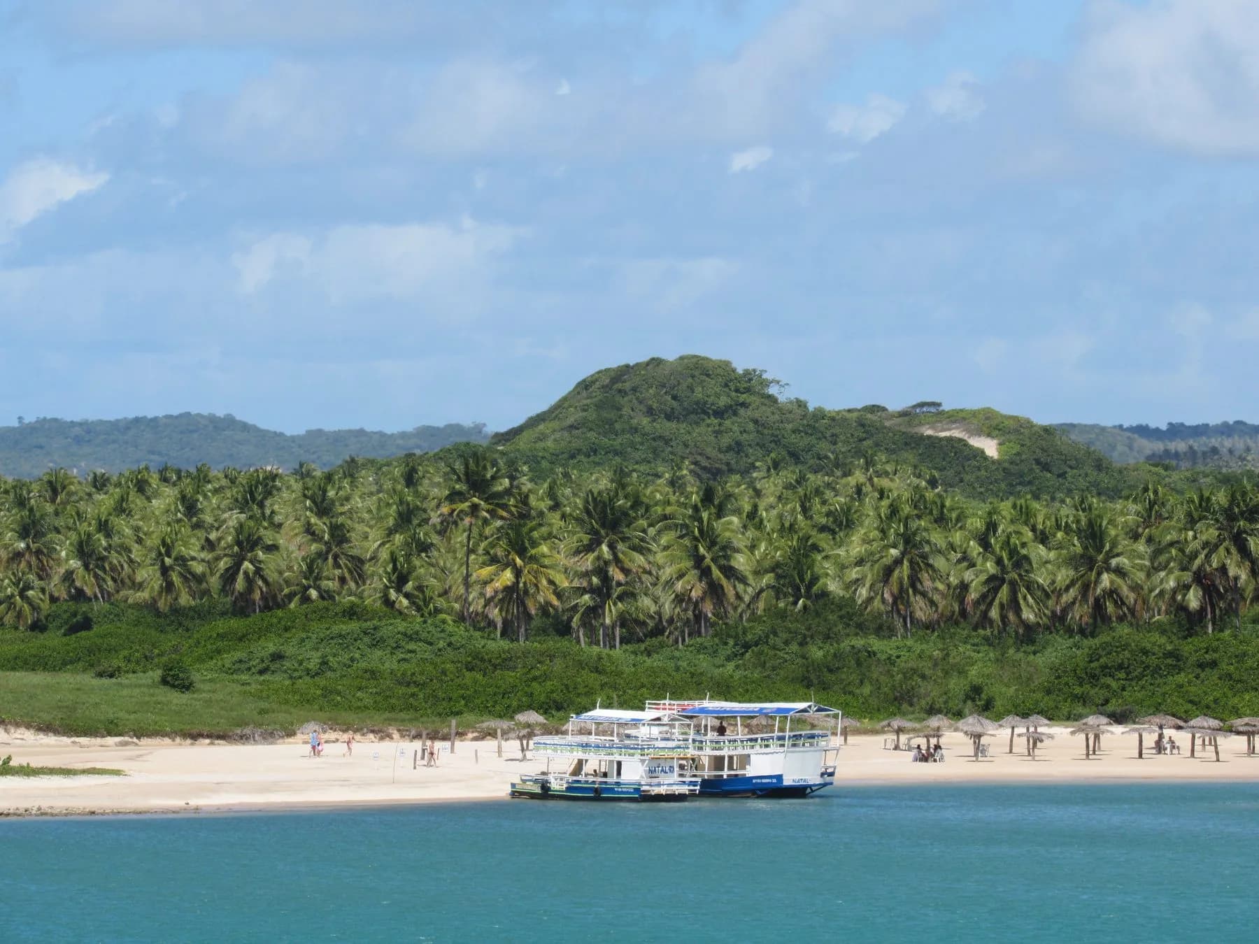Passeio para Barra do Cunhaú com barco diante da praia e de morro coberto por coqueiros
